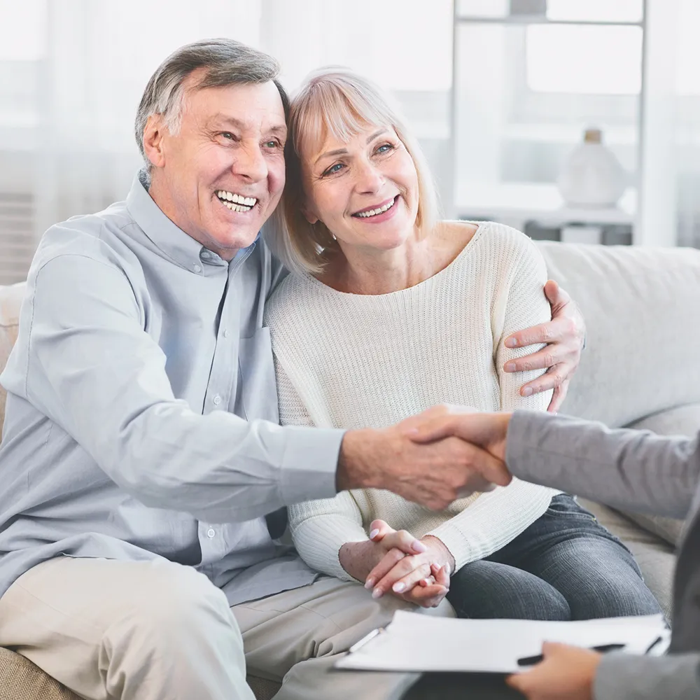 Casal sénior sorridente sentado num sofá, enquanto o homem aperta a mão de outra pessoa fora do enquadramento, transmitindo confiança e satisfação.