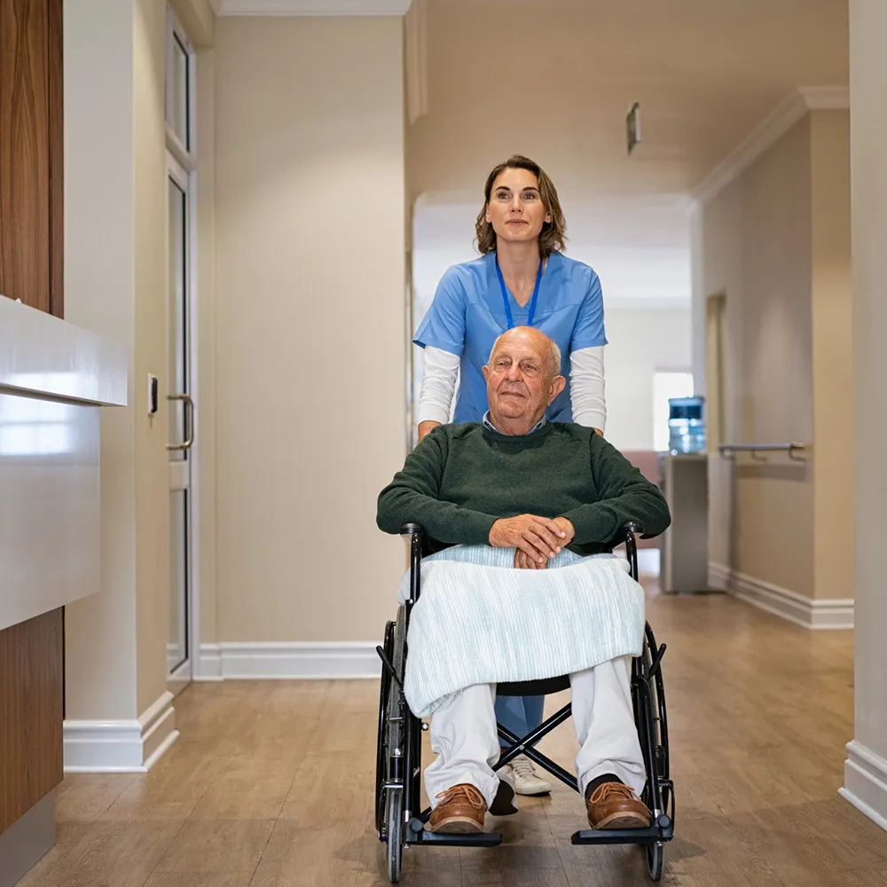 Profissional de saúde com uniforme azul a empurrar um homem idoso sentado numa cadeira de rodas, num corredor interior claro e espaçoso.